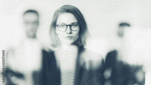 Woman with glasses stands in front of blurred figures in a bright room during a meeting at the office