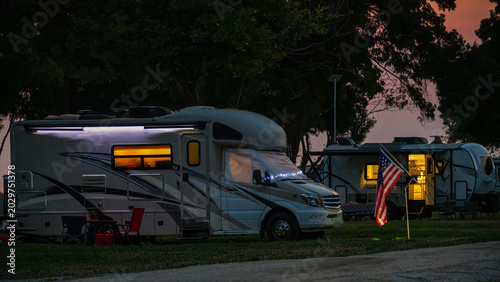 Two RVs are parked at a campsite during sunset, with lights glowing warmly inside. An American flag waves in the gentle evening breeze.