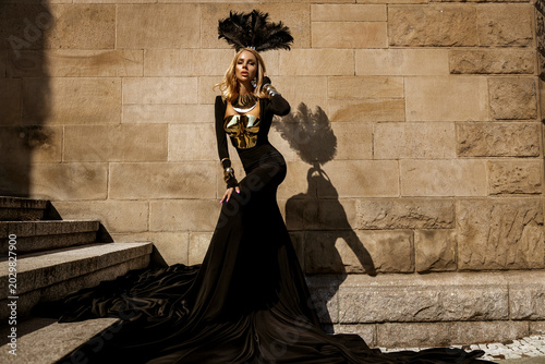 Fotografija Majestic female model in avant-garde flowing black gown with metallic gold armor and elaborate feather headdress posing against a textured stone wall in a high-fashion fantasy editorial