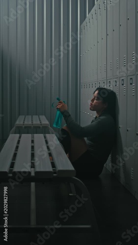 Young Sporty Woman Sitting on Floor, Leaning Against Lockers in Dressing Room. Girl Tired and Stares Blankly, Reflecting on Training. Burnout, Mental Challenge of Professional Sports. Vertical Shot.