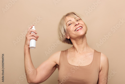 Quadro su tela Smiling mature woman with short blonde hair sprays facial mist on her face in a beige tank top against neutral background