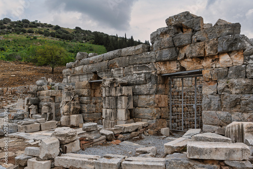 Ancient Ephesus ruins in Selcuk, Turkey. Historic stone walls, scattered blocks, and a metal gate against a green hillside under a cloudy sky, showcasing this UNESCO World Heritage site