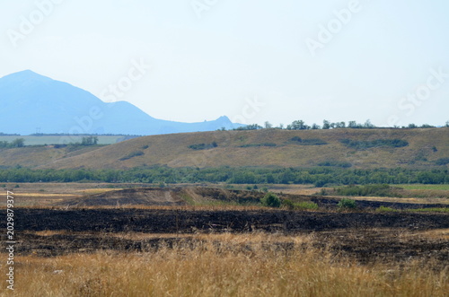 A beautiful landscape featuring a blue mountain peak rising behind a golden plateau and green agricultural fields. Clear summer day in the countryside with vast horizons and nature views