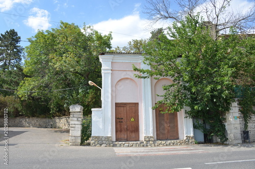 A small pink building with classical white arches and wooden doors located along an asphalt road. Lush green trees and a blue sky with clouds create a peaceful urban scene in summer day