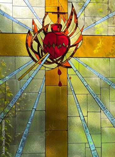 Detailed stained glass artwork depicting the Sacred Heart of Jesus and a golden cross inside a Catholic church. The vibrant colors, radiant light, and sacred symbolism create a powerful image of faith