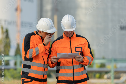 Fotografía Two workers in orange safety gear and helmets review documents outdoors at an industrial site