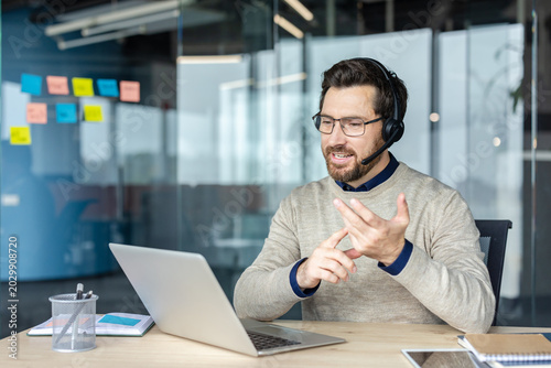 Businessman wearing a headset and spectacles using a laptop, having a video conference, and gesturing with his hands while communicating from a modern glass-walled office