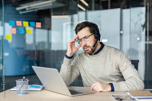 Photos Man in headset at office desk holding his head in pain, stressed and fatigued du