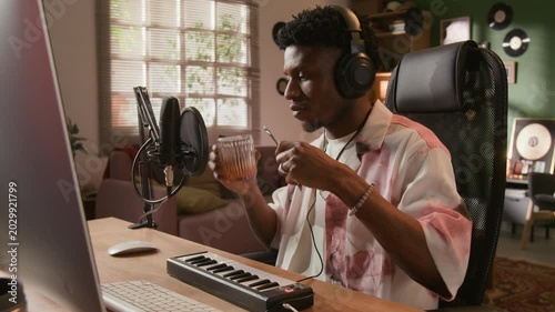 Medium shot of young adult African American music producer with headphones on recording metallic glass clinks in home based studio while creating experimental sound design