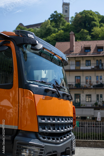 Bright orange construction truck parked in Ljubljana city center with castle tower in background during sunny summer day and urban European atmosphere