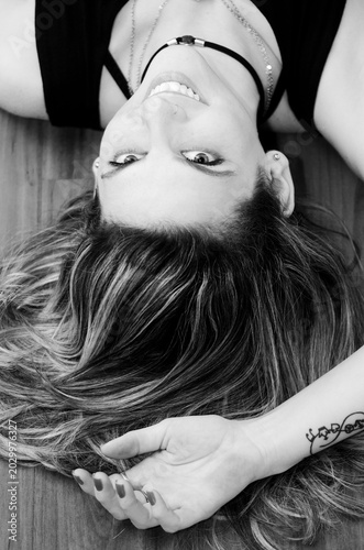 Black and white portrait of a woman lying down,photographed from above,with her face inverted in relation to the frame.The wide smile,loose hair,and accessories create a contemporary and relaxed image