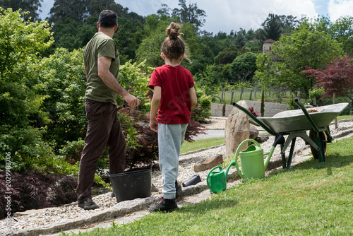 Schilderij op canvas A man and a child stand together in a scenic backyard garden next to landscaping tools, working on an outdoor family project