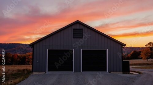 Modern garage building with dark grey metal siding and black doors sits against a vivid sunset sky. Hills and autumn trees create a scenic rural landscape background.