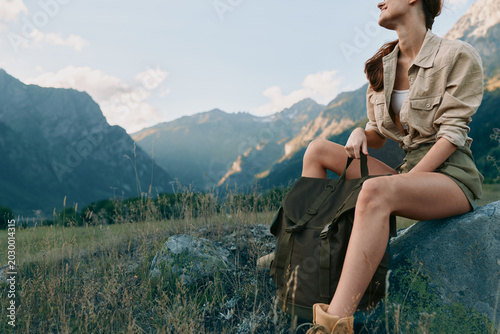 Photos Young woman on a hike rests on a rock, wearing a casual jacket and shorts, backpack beside, overlooking rugged mountains and a grassy meadow in a peaceful outdoor scene