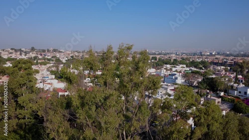 Aerial tilt up shot revealing Aqueduct of Queretaro rising above green trees and residential cityscape in Santiago de Queretaro Mexico during sunny daytime with urban skyline and historic arches