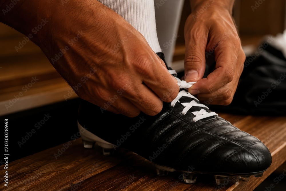 Fototapeta premium Athlete hands tying laces of black soccer cleats on a wooden bench in changing room