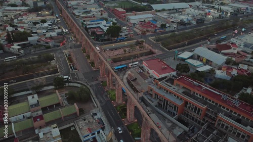 Drone tilt down cinematic view of Aqueduct of Queretaro surrounded by residential neighborhood and moving cars in Santiago de Queretaro Mexico at sunset