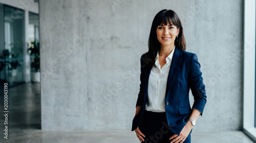 Confident businesswoman in navy blazer smiling directly at the camera against a modern concrete wall, conveying professionalism and success.