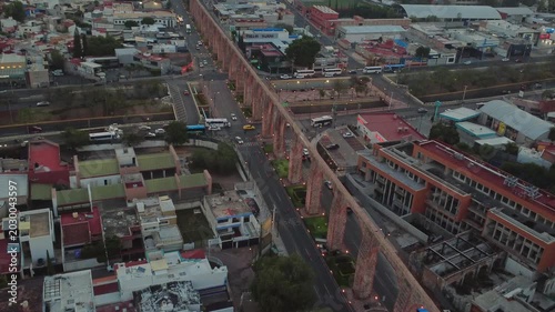 Dolly in aerial view approaching road crossing beneath Aqueduct of Queretaro with moving traffic and urban architecture in Santiago de Queretaro Mexico at dusk