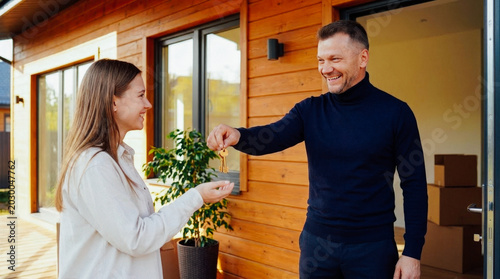 Happy couple receiving house keys from real estate agent at modern wooden house exterior on a sunny day, signifying new homeownership.