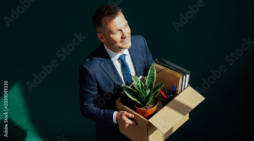Smiling middle-aged businessman carrying moving box with plant and files, conveying a sense of new beginnings and career transition.