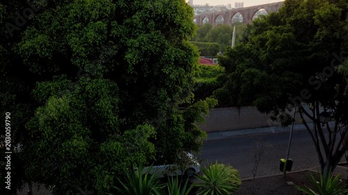 Tilt up drone shot from dense trees revealing Aqueduct of Queretaro and expanding urban skyline in Santiago de Queretaro Mexico during warm sunset light cinematic panorama