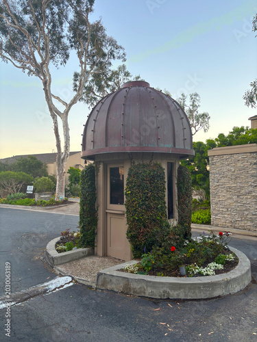 Small domed guardhouse or decorative kiosk surrounded by greenery and flowers in a landscaped residential community. Unique round architectural structure with metal dome roof, ivy-covered walls