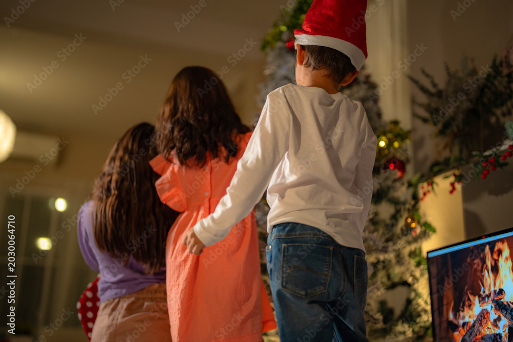 Obraz premium Three happy children celebrate Christmas at home, exchanging gifts near a beautifully decorated tree with cozy fireplace ambiance, enjoying holiday warmth, friendship, and family time together.