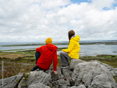 Father in red coat and daughter in yellow travel jacket are on top of a rough stone in a mountains, stunning nature scene with ocean and cloudy sky in the background. Divorced family concept. Travel.