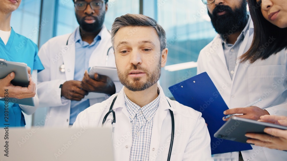 Obraz premium Head doctor working on laptop, surrounded by diverse medical team holding clipboards and tablets. Healthcare professionals focusing on shared data during team consultation. Close up of man.