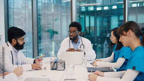 African American male doctor speaking during team meeting, surrounded by Indian man, Asian woman, and Caucasian woman taking notes. Multicultural medical team collaborating at conference table.