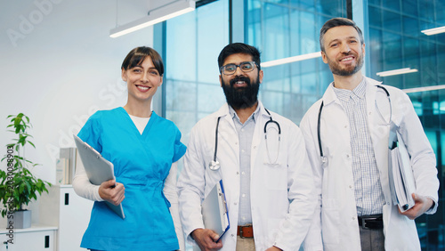 Portrait of group of young diverse doctors standing together in modern hospital office smiling. Confident medical team posing for photo. Female nurse and two male physicians holding clipboards. Indoor
