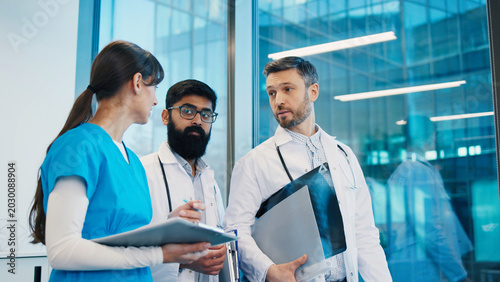 Female nurse and two male doctors examining chest X-ray by large glass window, holding medical documents and analyzing results. Diverse team discussing diagnosis in bright clinical office.