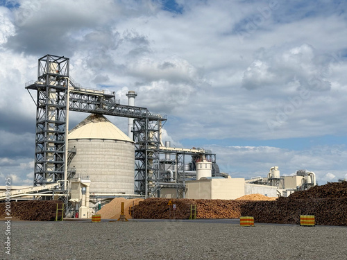 Industrial wood processing plant with large silos and timber stacks under cloudy sky