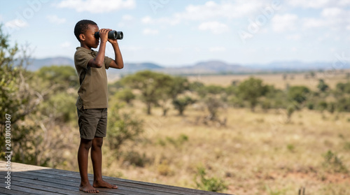 African boy on safari looking through binoculars at the savanna. Child on an adventure trip watching wildlife in Africa. Travel and tourism concept with copy space