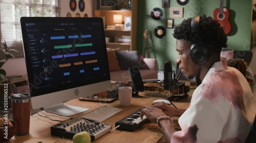 Side view of focused Black male music producer with headphones on playing MIDI keyboard while looking at DAW screen at home production setup