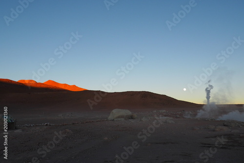 Stunning sunrise landscape at El Tatio geysers in Bolivia with steam columns rising against the moon, red volcanic mountains, and a clear blue sky, capturing an iconic South American travel destinatio