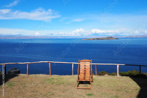 Empty wooden deck chair looking out over the deep blue waters of Lake Titicaca and the snow-capped Andes mountains under a sunny sky in Isla del Sol, Bolivia, perfect for scenic travel concepts.