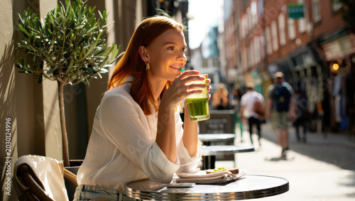 Young woman drinking green juice at outdoor cafe sunny city street healthy lifestyle wellness nutrition beauty glowing skin portrait