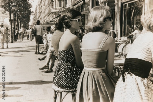 Stylish women in vintage dresses and sunglasses enjoying a sunny day at an outdoor Parisian cafe terrace.