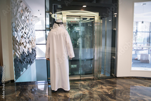Foto Man in traditional white robe stands in front of glass elevator in modern buildi