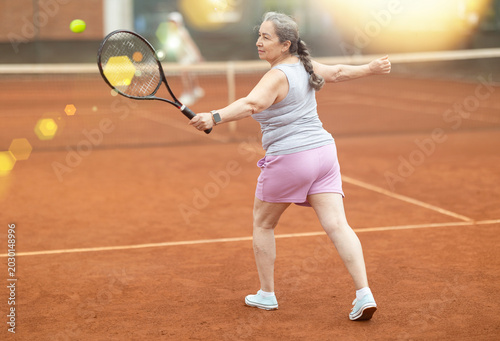 Fotografie Rear view of enthusiastic sporty senior woman, leading active healthy lifestyle, playing tennis with female opponent behind net in background on outdoor court on summer day
