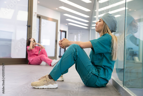 Two female healthcare professionals in scrubs and surgical caps sit on a hospital floor, appearing stressed and fatigued from occupational burnout.