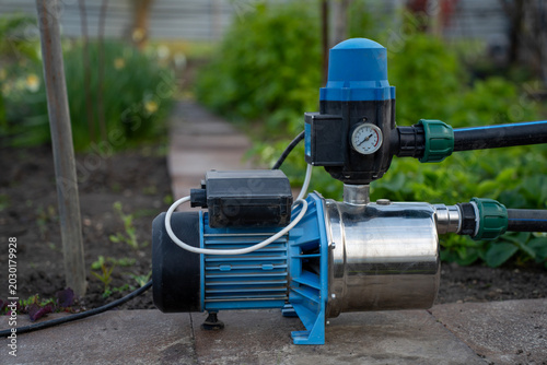 Fotografie Surface water pump with automatic pressure control switch and gauge installed on a garden path tile, connected to black irrigation pipes with green fittings