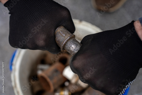 Photos Hands in black protective gloves trying to unscrew or assemble an old rusty metal threaded pipe joint over a bucket containing plumbing scrap