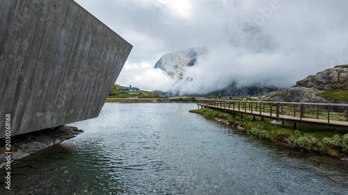 Andalsnes, Norway - 23 June 2024: View of the Trollstigen visitor center and wooden walkway by a mountain lake under a cloudy sky.