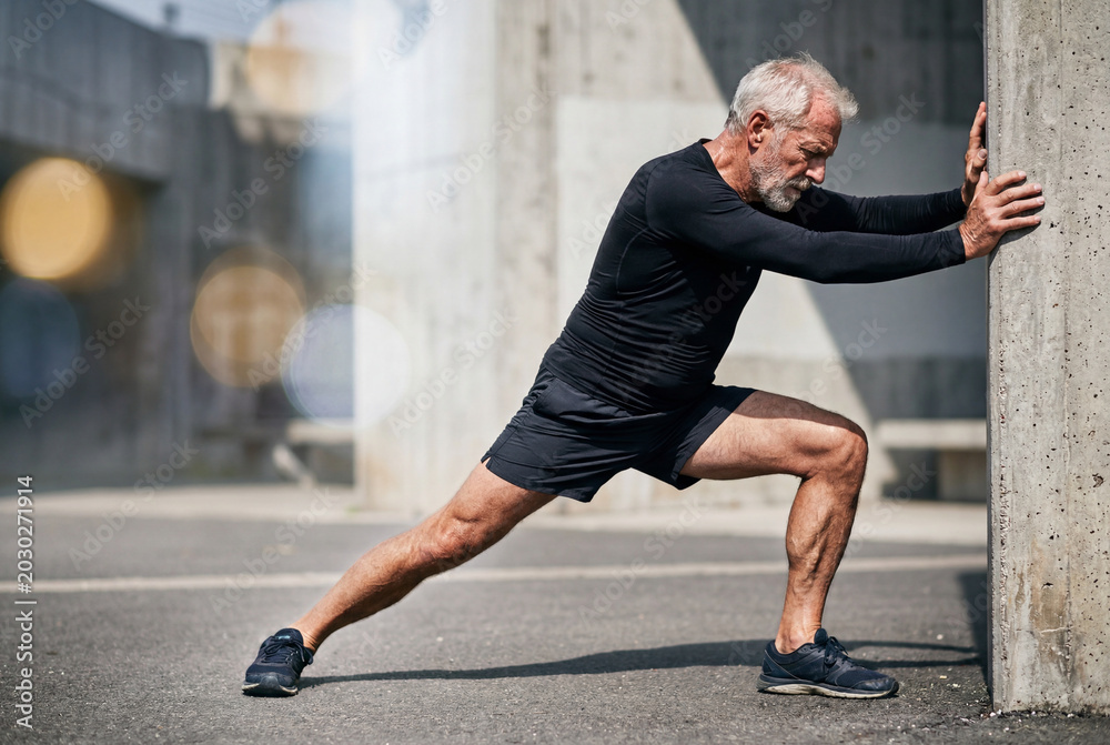 Fototapeta premium Senior man in dark athletic clothing stretching his leg muscles against a massive concrete wall