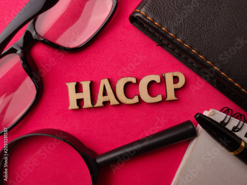 Wooden letters spelling HACCP are arranged on a bright pink surface alongside glasses, a notebook, pen, and magnifying glass