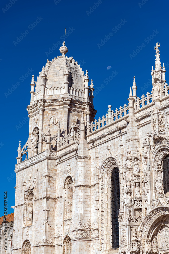 Fototapeta premium Lisbon, Portugal - October 11, 2025 - Close-up detail of the intricate Manueline style architecture and spires of the Jeronimos Monastery against a blue sky with the moon.