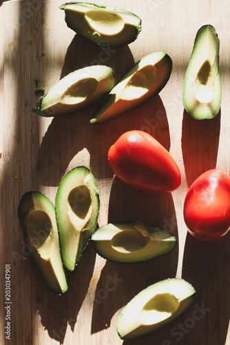Fresh green avocado sliced and ready to eat along with whole organic roma tomatoes on a cutting board. Delicious healthy, nutritious food.